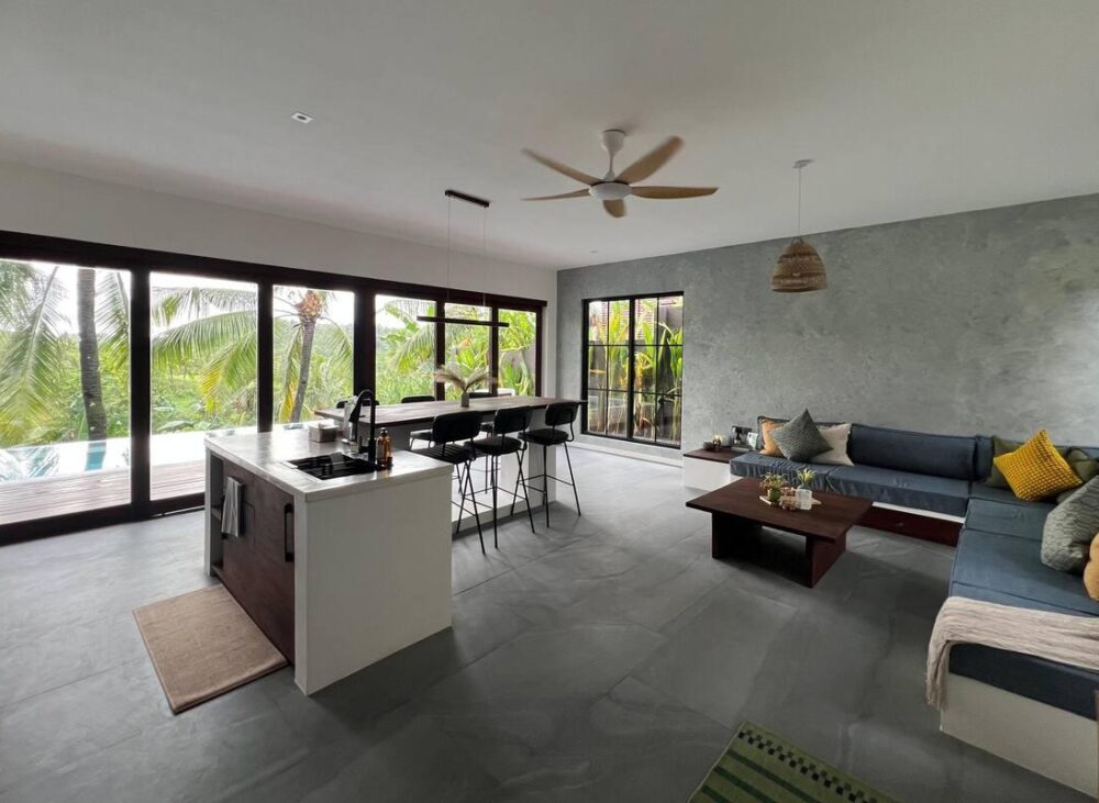 Modern open-plan living room, dining area, and kitchen in a contemporary Kerala home with abundant natural light.