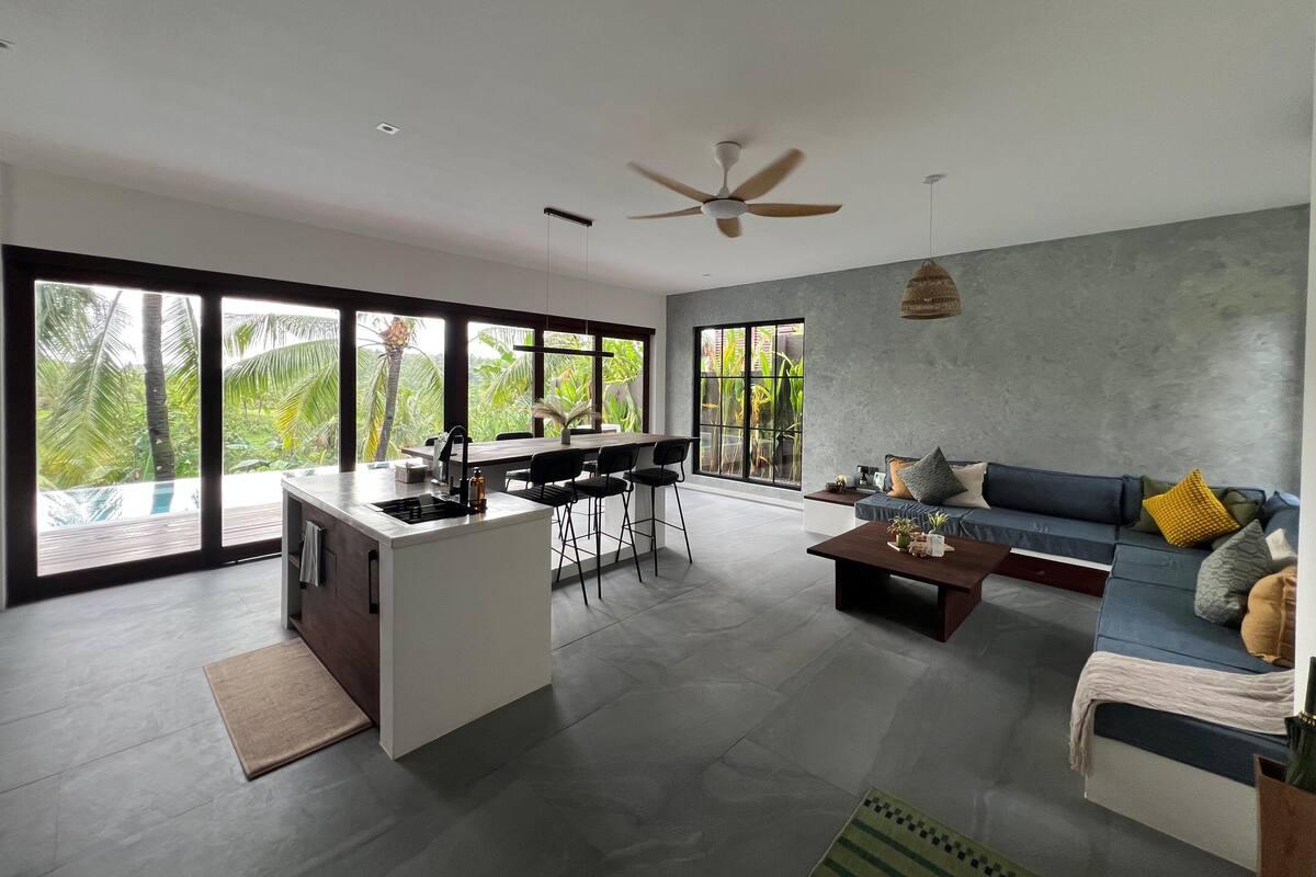 Modern open-plan living room, dining area, and kitchen in a contemporary Kerala home with abundant natural light.