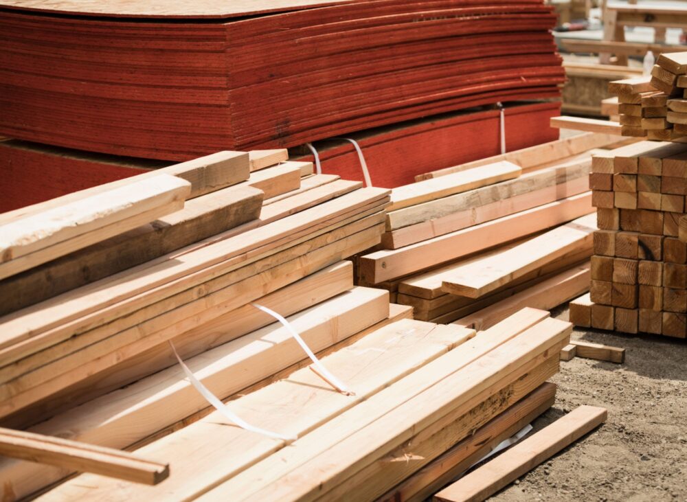 A side-by-side comparison of building materials in a warehouse, featuring a neat stack of thick, light-colored solid wood timber beams in the foreground and a large pile of thin, reddish-brown plywood sheets in the background.