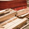 A side-by-side comparison of building materials in a warehouse, featuring a neat stack of thick, light-colored solid wood timber beams in the foreground and a large pile of thin, reddish-brown plywood sheets in the background.