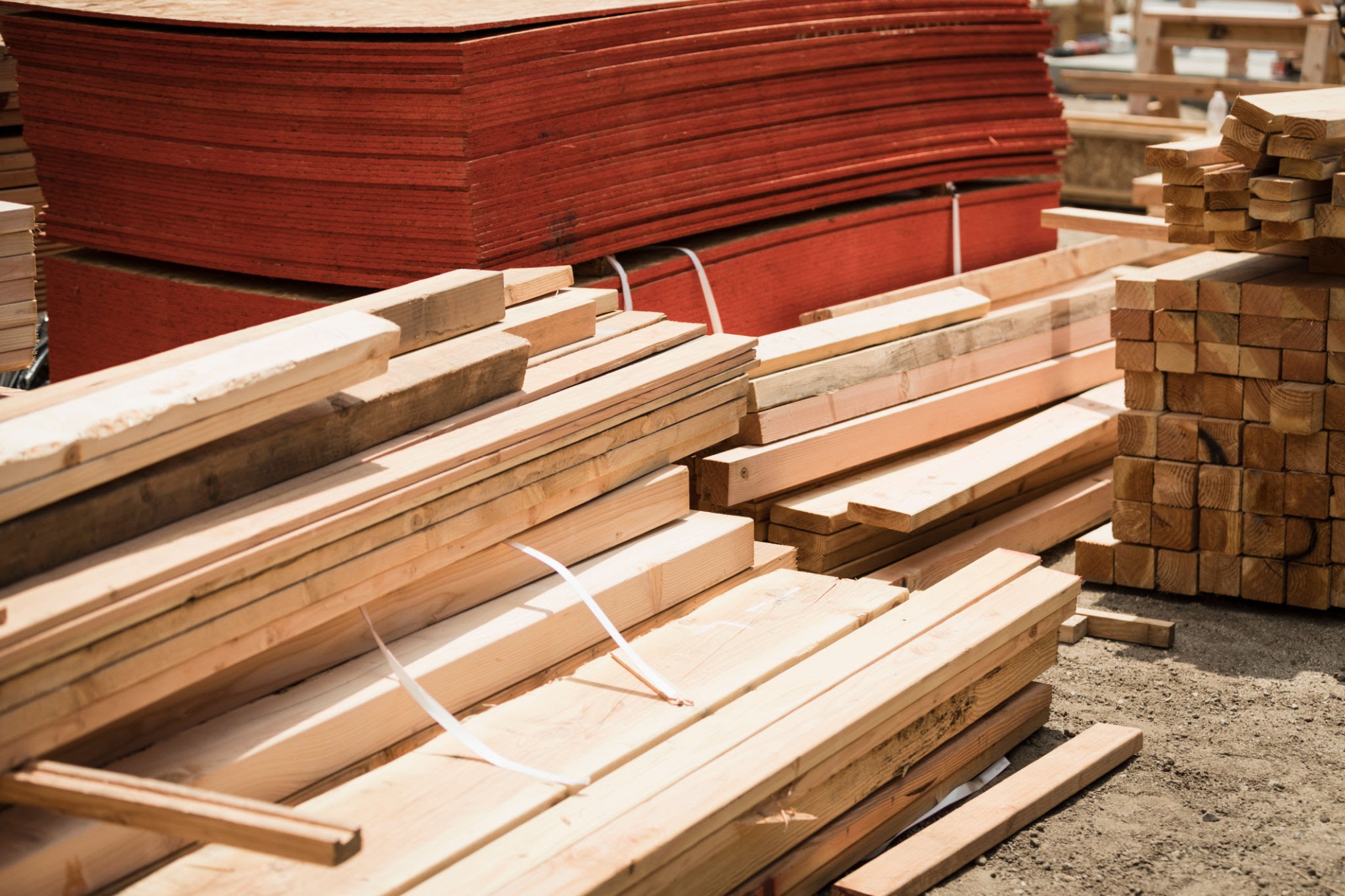 A side-by-side comparison of building materials in a warehouse, featuring a neat stack of thick, light-colored solid wood timber beams in the foreground and a large pile of thin, reddish-brown plywood sheets in the background.